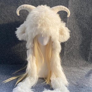 Op de afbeelding: Een witte muts van imitatiebont met twee gebogen, getextureerde hoorns aan de bovenkant en lange, ruige oorflappen. De muts wordt getoond op een etalagepophoofd met lang, steil blond haar dat onder het pluizige materiaal vandaan komt.