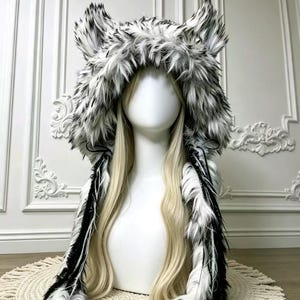 May include: A faux fur hood featuring a black and white wolf-like pattern with prominent pointed ears. The hood includes long, dangling side panels with black and white fur trim, designed to frame the face and drape down the shoulders.