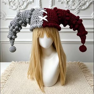 May include: A two-toned crochet jester hat featuring a split design with one side in light grey and the other in deep burgundy. The hat has long, curved, textured horns with bobble details, finished with pom-poms and a white ribbon lace-up.
