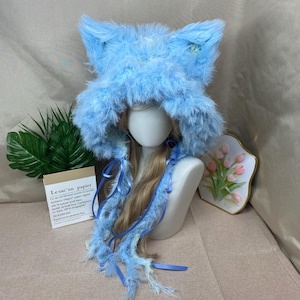 May include: A light blue, fluffy cat ear hat with long, flowing accents and ribbons. The hat is displayed on a mannequin head with long blonde hair. The hat's design includes pointed ears and a soft, textured appearance.
