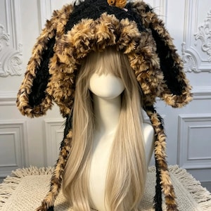 May include: A plush hat featuring long, floppy ears with a black and brown faux fur texture. The hat has a small, light brown stuffed animal attached to the top center. Two long, fur-lined ear flaps extend downward from the sides of the headpiece.