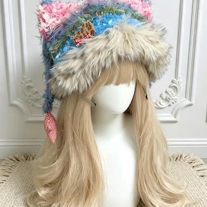 Può includere: Un cappello con orecchie da gatto in filato testurizzato blu e rosa con un bordo spesso in pelliccia sintetica color crema. Il cappello è decorato con una piccola toppa a forma di cuore leopardato e un ciondolo a cuore a quadretti rossi e bianchi.