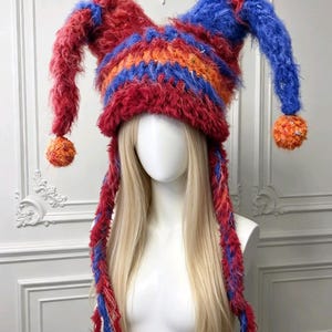 Può includere: Un cappello in stile giullare realizzato in lana soffice, caratterizzato da due lunghe corna appuntite sulla parte superiore, ciascuna terminante con un pompon testurizzato arancione e rosso. Il cappello presenta strisce orizzontali di lana rossa, blu e arancione.