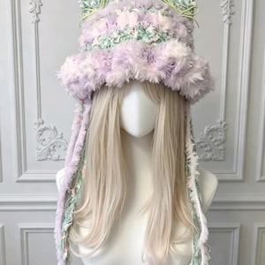 May include: A fuzzy bucket hat featuring cat-like ears, crafted from a textured yarn blend in shades of pastel lavender, mint green, and white. The hat includes long, dangling side ties made of matching variegated yarn and soft, plush fibers.