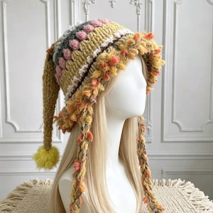 May include: A multi-textured, long-tailed crochet hat featuring horizontal stripes in mustard yellow, soft pink, and grey. The brim is adorned with a thick, shaggy fringe of orange and yellow yarn, and the long tail ends in a bright yellow pom-pom.