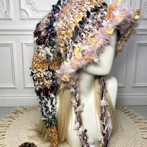 May include: A long, pointed crochet hat featuring a multi-textured blend of yarn in shades of purple, yellow, white, and black. The hat has a wide, fuzzy brim, long braided side ties, and a large, round pom-pom at the tip of the tail.