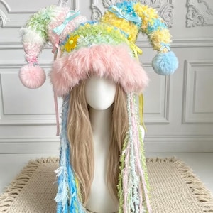 May include: A multi-coloured jester hat featuring a fuzzy pink brim, two long pointed sections with light blue pom-poms, and long hanging fringe strands in pastel shades of blue, green, yellow, and pink, accented with small satin ribbons and a yellow flower.