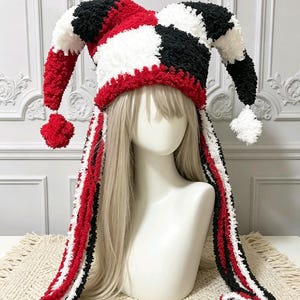 May include: A jester hat crafted from textured yarn in a colour-blocked pattern of red, white, and black. The hat features two long, dangling side panels and two pointed top sections, each finished with a round pom-pom at the end of the points.