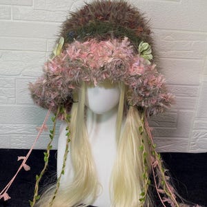 May include: A textured bucket hat featuring a dark brown and olive green fuzzy crown with a wide, shaggy pink brim. The hat is adorned with small pale green fabric flowers and long, dangling pink braided cords intertwined with green leaf vines.