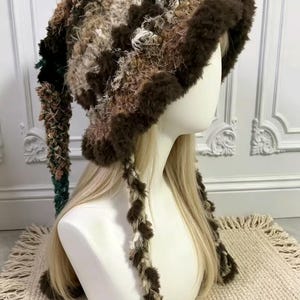 May include: A textured bucket hat crafted from a mix of brown, beige, and cream yarns. The hat features a fuzzy, dark brown trim along the brim and two long, braided side ties that hang down, incorporating shades of brown, cream, and dark green.