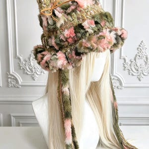 May include: A fuzzy bucket hat featuring a multi-colored pattern of olive green, soft pink, and cream fibers. The hat has two pointed cat-like ears at the top, each accented with a small tan leather bow, and two long, dangling braided side ties.