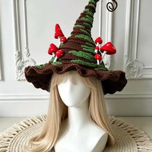 Puede incluir: Un sombrero de ganchillo alto y puntiagudo en color marrón oscuro con detalles verdes texturizados y varias setas pequeñas de color rojo brillante con puntos blancos adheridas al exterior. Tiene un ala ancha y ondulada y una punta rizada.