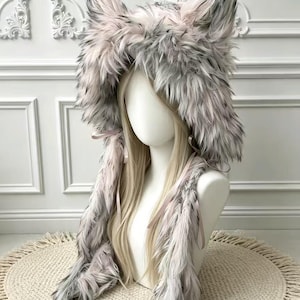 May include: A faux fur hood featuring tall, pointed animal ears in shades of grey, white, and soft pink. The hood is lined with long, shaggy fur and includes two long, dangling side panels that hang down, secured with thin, light pink ribbon ties.