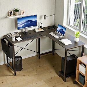 Könnte beinhalten: Ein L-förmiger Computertisch mit dunkler Holzmaserung und schwarzen Metallbeinen. Der Schreibtisch hat einen Computermonitor, eine Tastatur, eine Maus und einen Laptop. Ein schwarzer Mülleimer und ein schwarzer Zeitschriftenhalter befinden sich auf der linken Seite.