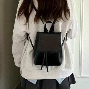 Può includere: Zaino nero con patta quadrata e chiusura con coulisse. Il piccolo zaino ha un manico superiore e spallacci regolabili. La borsa è indossata sopra una felpa grigio chiaro e una gonna a pieghe nera.