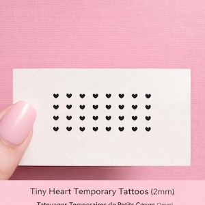 May include: A sheet of temporary tattoos featuring a grid of small black heart shapes, each 2mm in size. The sheet is held against a pink background. Text at the bottom reads "Tiny Heart Temporary Tattoos (2mm)".