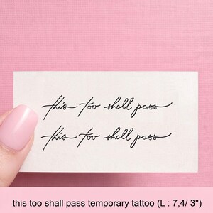 Peut inclure: Un tatouage temporaire avec l'inscription "this too shall pass" en écriture noire élégante. Le tatouage est sur une petite carte rectangulaire. Le fond est rose clair.