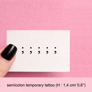 Peut inclure: Un tatouage temporaire blanc avec cinq symboles point-virgule noirs. La feuille est posée sur un fond rose. Le texte en bas indique "semicolon temporary tattoo (H : 1,4 cm/ 0,6")".