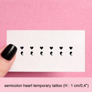 Peut inclure: Une carte rectangulaire blanche avec six tatouages temporaires en forme de cœur-point-virgule noirs. Les tatouages sont disposés en ligne horizontale. La carte est tenue sur un fond rose. Le texte "semicolon heart temporary tattoo (H : 1 cm/0,4")" est en bas.