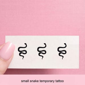 Puede incluir: Una hoja de tatuajes temporales con tres pequeños diseños de serpientes negras. Los tatuajes se muestran en una tarjeta rectangular blanca sobre un fondo rosa. El texto "small snake temporary tattoo" está impreso debajo.