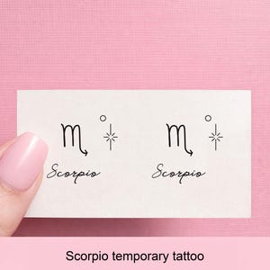 Puede incluir: Una hoja de tatuaje temporal con dos símbolos del zodiaco Escorpio negros y la palabra "Scorpio" en cursiva debajo. Cada símbolo está acompañado de una pequeña explosión de estrellas. El fondo es de color rosa claro.