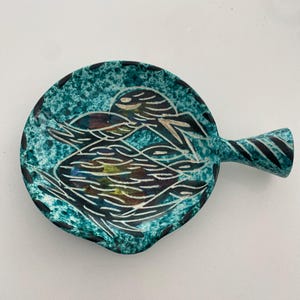 Peut inclure: Un plat en céramique turquoise avec une poignée, présentant une surface texturée et un motif blanc et noir. Le plat est de forme ronde avec une poignée, et le motif semble être un oiseau ou un poisson.