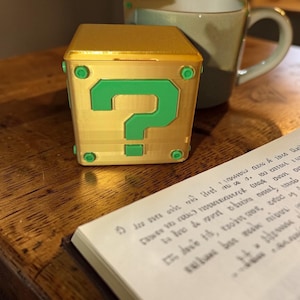 Peut inclure: Un cube doré avec un point d'interrogation vert, rappelant un objet de jeu vidéo. Le cube a des accents verts et se trouve près d'une tasse et d'un carnet.