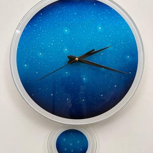 May include: A round wall clock with a blue and white galaxy design. The clock has black hands and a silver frame. A smaller, matching circular pendulum hangs below the clock.