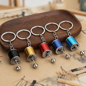 May include: Five miniature gear shift keyrings in black, gold, red, blue, and teal. Each keyring features a silver-coloured gear shift lever and a small, round ball at the end. The keyrings are displayed on a wooden tray.