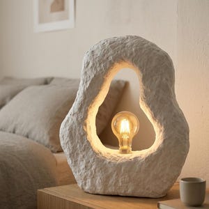 May include: An off-white, textured table lamp with an organic, open-centre design. The lamp features a warm, glowing lightbulb. It sits on a wooden bedside table, with a small cup and book. The lamp's unique shape adds a modern touch to the room.