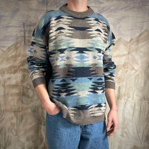 May include: A patterned crew neck sweater in blue, grey, and brown. The sweater has a ribbed collar, cuffs, and hem. The design features repeating geometric shapes and lines. The sweater is a classic fit.