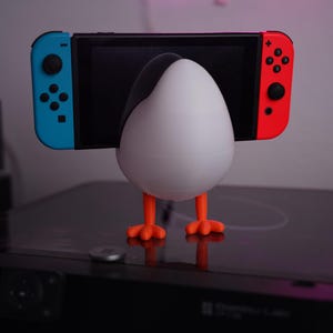 Può includere: Un supporto a forma di pinguino bianco e nero che tiene una Nintendo Switch. Il supporto ha zampe arancioni ed è progettato per sostenere la console di gioco. Lo Switch ha controller blu e rossi.