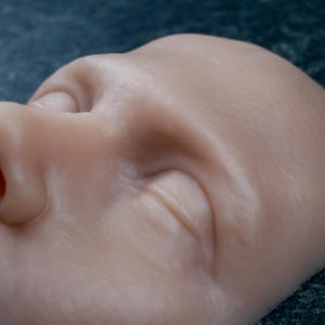 Può includere: Primo piano di una maschera facciale color carne pallido. La maschera presenta un occhio chiuso, un naso e parte della guancia. La superficie presenta trame e ombre sottili, che suggeriscono un volto umano realistico. Lo sfondo è verde scuro screziato.