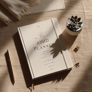 May include: Cream-colored ADHD planner with the text "ADHD PLANNER" in gold lettering. The planner has a matching elastic band and a ribbon bookmark. A gold pen, succulent plant, and paper clips are also visible.