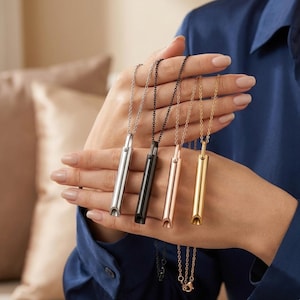 May include: Four cylindrical pendant necklaces hanging from delicate metal chains. The pendants are finished in silver, black, rose gold, and gold tones. Each pendant features a hollow, whistle-like opening at the bottom and a small bail at the top.