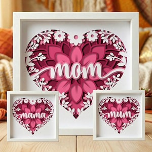 Könnte beinhalten: Ein Set aus drei gerahmten Papierkunstwerken. Das größte zeigt ein geschichtetes Herzdesign mit dem Wort "mom" in weißer Schrift. Die beiden anderen kleineren Rahmen zeigen "mama" und "mum". Die Kunst ist in Rosa- und Weißtönen gehalten.