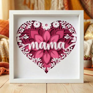 May include: A framed, layered paper art piece featuring a heart design. The heart is filled with pink and magenta layers, with the word "mama" in white script. White floral and leaf cutouts surround the heart, set against a white frame.