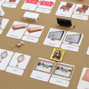 May include: A collection of educational flashcards featuring illustrations of furniture and home decor items like sofas, armchairs, coffee tables, televisions, clocks, and pictures, accompanied by miniature 3D models of a white table and a black television.