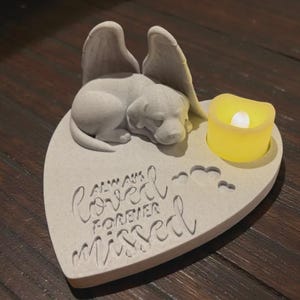 Op de afbeelding: Een hartvormig gedenkteken met een slapende puppy, engelen vleugels en een gele LED-kaars. De tekst "Always Loved, Forever Missed" is op het oppervlak gegraveerd. Het item is gemaakt van lichtgrijs materiaal.