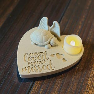 May include: A heart-shaped memorial candle holder with a turtle, angel wings, and the words "Always Loved, Forever Missed." The candle holder has a small, lit, yellow candle. The item is made of a light beige material.