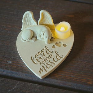 May include: A heart-shaped memorial with a sleeping dog figurine, angel wings, and a small candle. The text "Always Loved Forever Missed" is inscribed on the heart. The piece is made of a light-colored material.