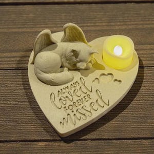 Op de afbeelding: Een hartvormig gedenkteken met een slapende kattenengel figuur en een kleine LED-kaars. De basis is gegraveerd met de woorden "Always Loved, Forever Missed". De kat heeft engelvleugels. De kleur is lichtbeige.