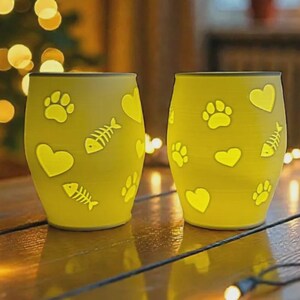 May include: Two yellow glass tumblers with paw prints, hearts, and fishbone designs. The tumblers are illuminated, creating a warm glow. The background features blurred lights, suggesting a festive or cozy setting.