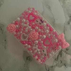 May include: A clear phone case decorated with pink and white pearls, hearts, and roses. The case features a pink bow and a cartoon character with a purple bow. The case is covered in small, decorative elements.