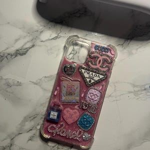 Puede incluir: Una funda de teléfono rosa brillante adornada con varios adornos inspirados en diseñadores. Incluye un frasco de perfume Chanel, el logotipo de Prada y la palabra "Chanel" en pedrería. También hay otros amuletos y el texto "Baby Bad Energy".