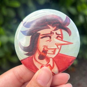 May include: A round button featuring a cartoon illustration of a man with dark hair, a long nose, and a red suit. The button has a glossy finish and a light green background.