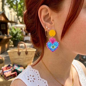 Pode incluir: Brincos coloridos em forma de coração. Cada brinco tem um círculo amarelo, um design de coração rosa e azul e uma corrente dourada. O design do coração apresenta um olho. Usados por uma pessoa com cabelo ruivo.