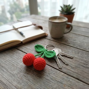 Op de afbeelding: Een rode gehaakte kersen sleutelhanger met groene bladeren en een zilveren sleutelring ligt op een houten tafel. Daarnaast liggen sleutels, een open notitieboek met pen en een mok koffie. Een kleine vetplant in een pot is ook zichtbaar.