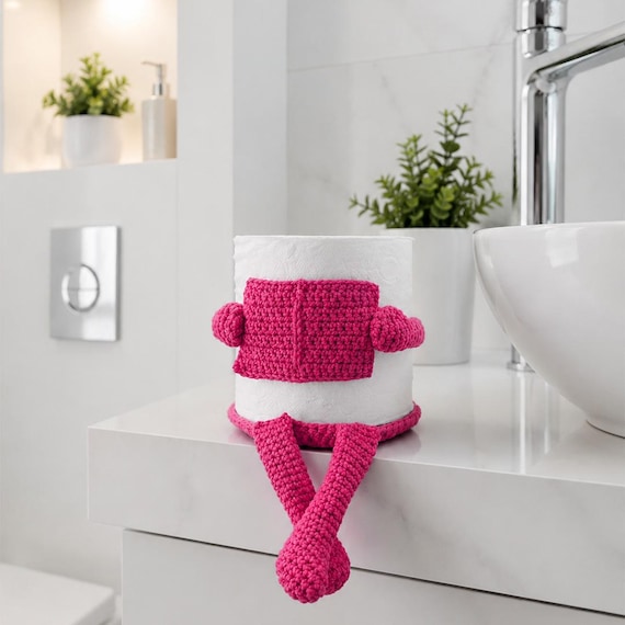 A hand-crocheted toilet paper cozy styled as a funny reading companion — the kind of quirky bathroom decor that guests will actually comment on.