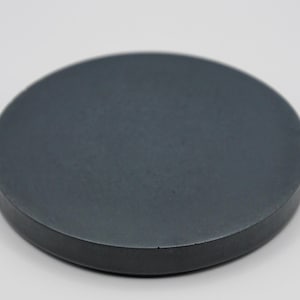 May include: A solid, round, dark gray coaster. The coaster is made of a smooth material and has a slightly raised edge. The object is a simple, minimalist design, suitable for use with cups and glasses.